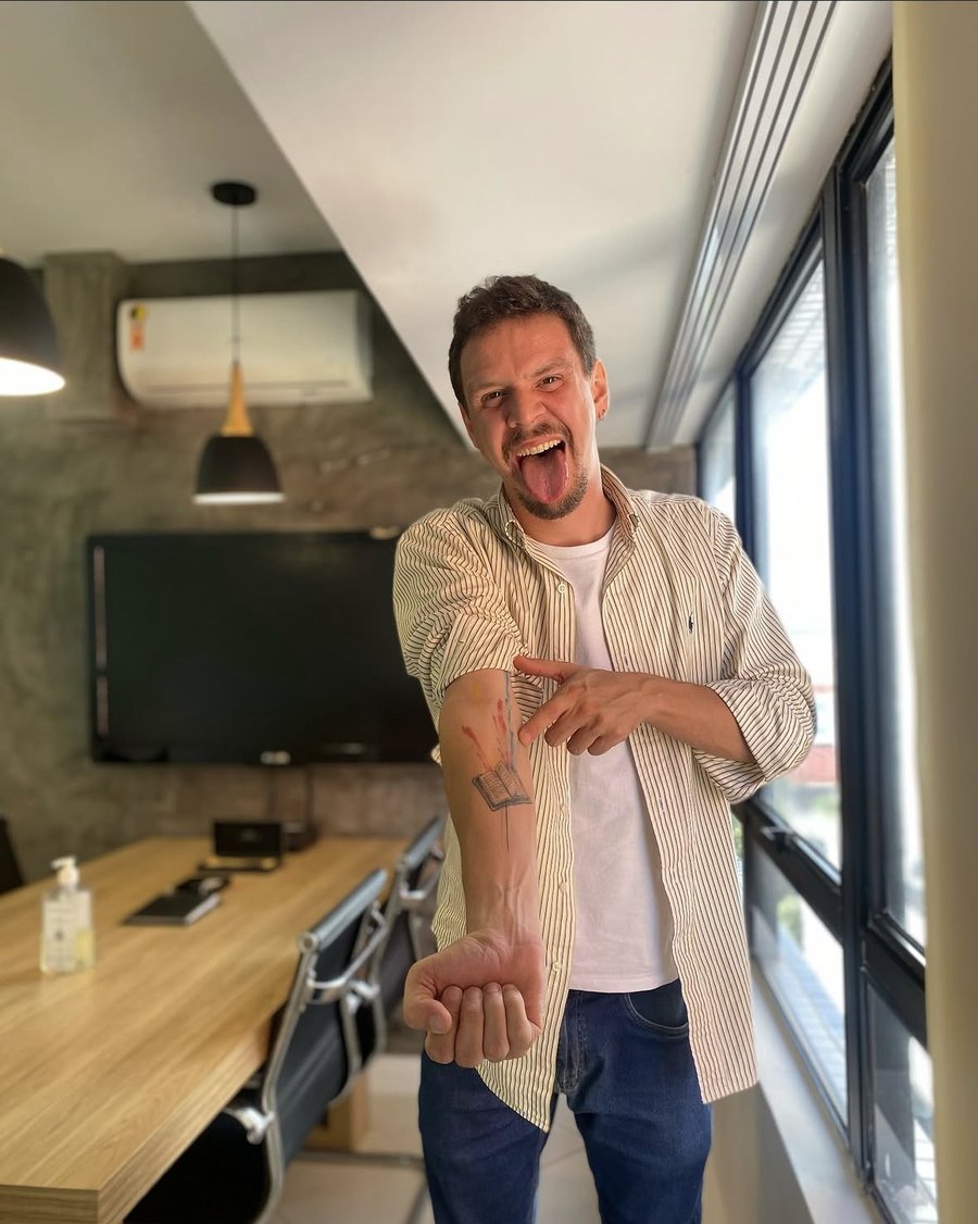 João Guilherme Magalhães no escritório, em clima descontraído, mostrando a tatuagem de um livro no antebraço